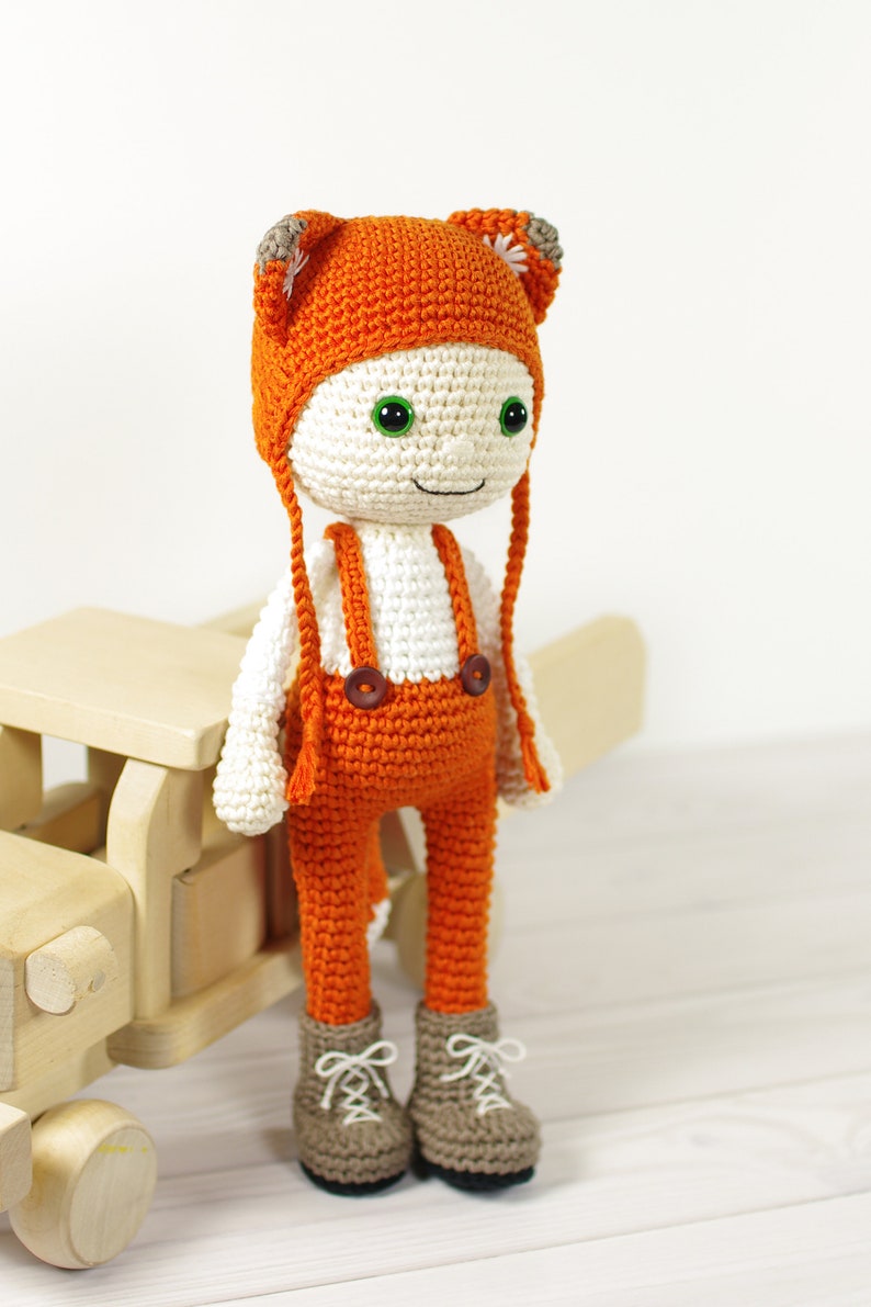 May include: A crocheted fox doll wearing orange overalls and a matching hat with fox ears. The doll has green eyes and a white face. The doll is standing next to a wooden toy.