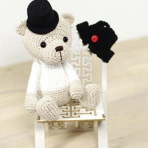 May include: A crocheted teddy bear wearing a black top hat and a black vest with a red button. The bear is sitting on a white wooden chair with a decorative pattern.
