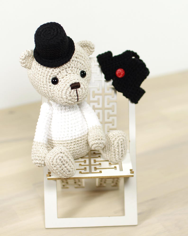 May include: A crocheted teddy bear wearing a black top hat and a black vest with a red button. The bear is sitting on a white wooden chair with a decorative pattern.