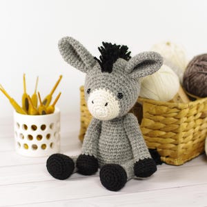 May include: A handmade gray crochet donkey with black hooves and a black mane sits on a white surface. The donkey has a white muzzle and large ears. A white ceramic container with yellow crochet hooks and a basket of yarn balls are in the background.