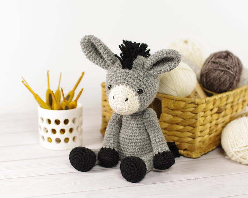 May include: A handmade gray crochet donkey with black hooves and a black mane sits on a white surface. The donkey has a white muzzle and large ears. A white ceramic container with yellow crochet hooks and a basket of yarn balls are in the background.