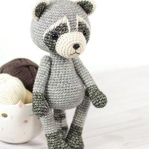 May include: A crocheted raccoon toy with gray and black fur. The toy has a white nose and black eyes. The toy is sitting on a white wooden surface.