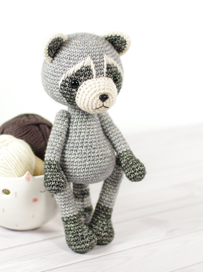 May include: A crocheted raccoon toy with gray and black fur. The toy has a white nose and black eyes. The toy is sitting on a white wooden surface.