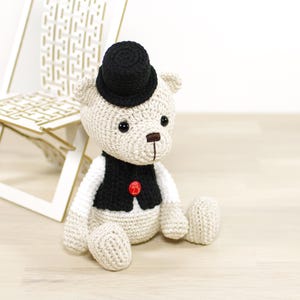 May include: A crocheted teddy bear wearing a black top hat and a black vest with a red button. The bear is sitting on a light wood surface.