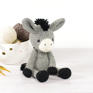 May include: A crocheted donkey toy in shades of grey, white, and black. The donkey has a grey body, black hooves and mane, and a white muzzle. It is sitting on a white surface. A bowl of yarn is in the background.