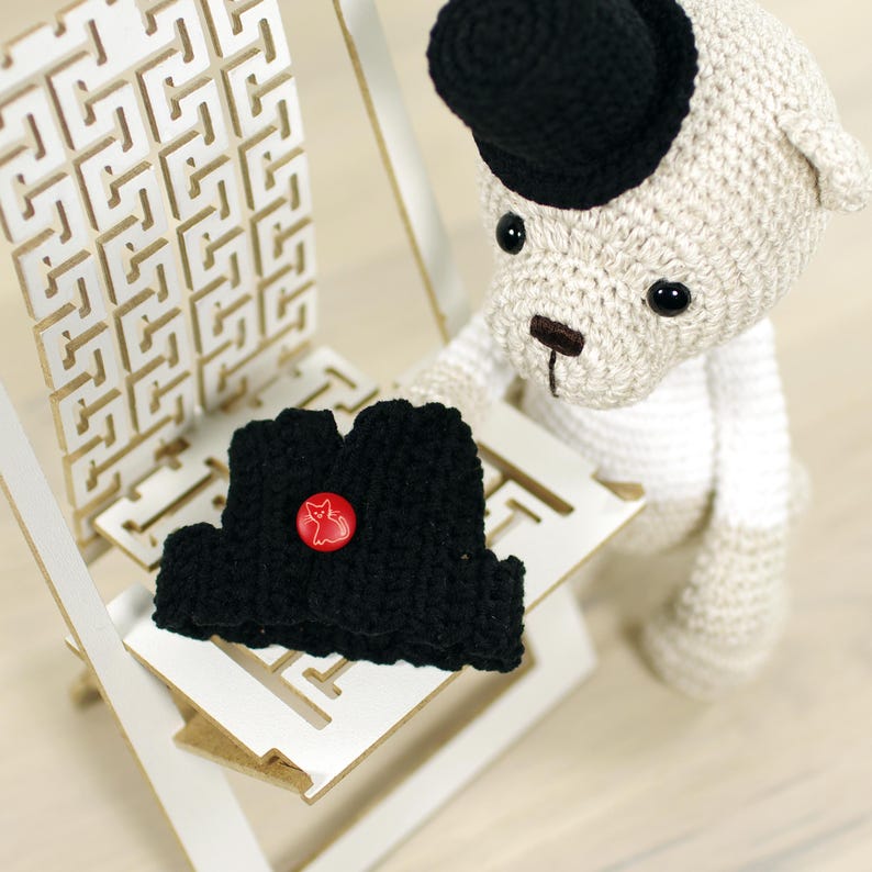 May include: A white crocheted teddy bear wearing a black top hat and a black crocheted vest with a red button. The bear is sitting on a white wooden chair.
