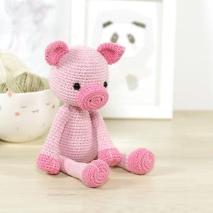 Puede incluir: Un juguete de cerdo de crochet rosa con ojos negros y un hocico rosa. El cerdo está sentado sobre una superficie de madera.