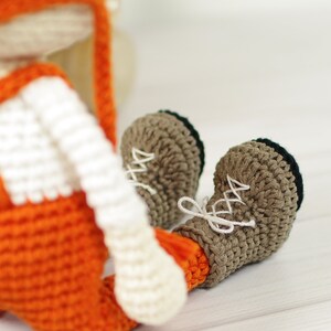 May include: A crocheted fox toy wearing orange overalls and brown boots. The fox has white fur and a long, pointed nose.