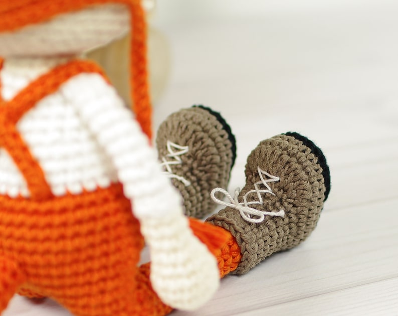 May include: A crocheted fox toy wearing orange overalls and brown boots. The fox has white fur and a long, pointed nose.