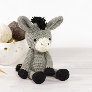 May include: A handmade crochet donkey toy in shades of gray, black, and white. The donkey has a black mane and hooves, with a white muzzle. A white ceramic bowl filled with yarn and two yellow crochet hooks are in the background.