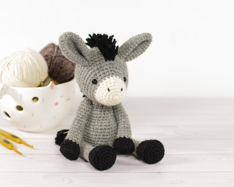 May include: A handmade crochet donkey toy in shades of gray, black, and white. The donkey has a black mane and hooves, with a white muzzle. A white ceramic bowl filled with yarn and two yellow crochet hooks are in the background.