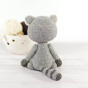 May include: A gray crocheted cat toy sitting on a white wooden surface. The cat has a striped tail and white accents on its ears.