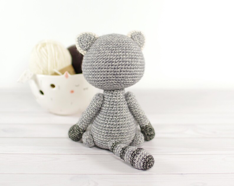 May include: A gray crocheted cat toy sitting on a white wooden surface. The cat has a striped tail and white accents on its ears.