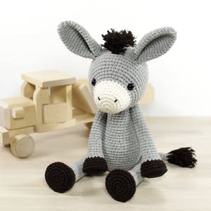 May include: A grey crocheted donkey plush toy with brown accents, sitting on a wooden floor. The donkey has a white face with black eyes and a brown mane.
