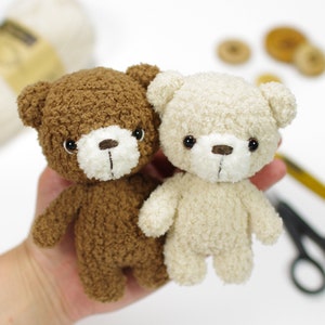 May include: Two plush teddy bears, one brown and one cream, with black eyes and brown noses. The bears are standing on a hand.