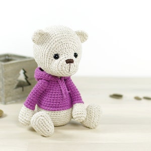 May include: A crocheted teddy bear wearing a pink hooded sweater. The bear is sitting on a light wood surface.