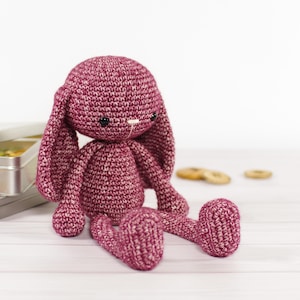 Puede incluir: Un juguete de conejo de crochet rosa con orejas largas y caídas. El conejo está sentado sobre una superficie de madera blanca.