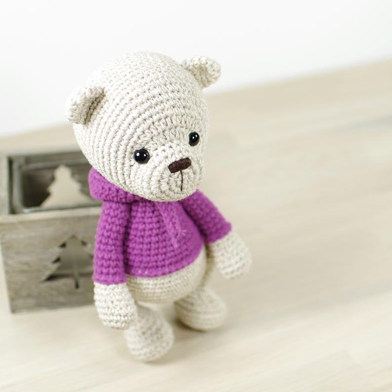 K&ouml;nnte beinhalten: Ein handgeh&auml;kelter Teddyb&auml;r mit hellbeigem K&ouml;rper und Kopf, der einen lila Kapuzenpullover tr&auml;gt. Der B&auml;r hat schwarze Augen und eine braune Nase. Das Spielzeug befindet sich in der N&auml;he einer Holzkiste mit einem Weihnachtsbaum-Ausschnitt.
