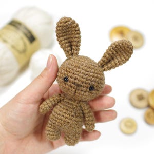 Könnte beinhalten: Ein kleiner brauner Häkel-Hasen mit schwarzen Augen. Der Hase wird in der Hand einer Person gehalten.