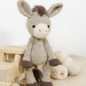 May include: A crocheted donkey soft toy made from light beige yarn, featuring a cream-coloured muzzle, dark brown hooves, and a dark brown tuft of hair on its head and tail. The donkey has two large ears and small black eyes, standing upright.