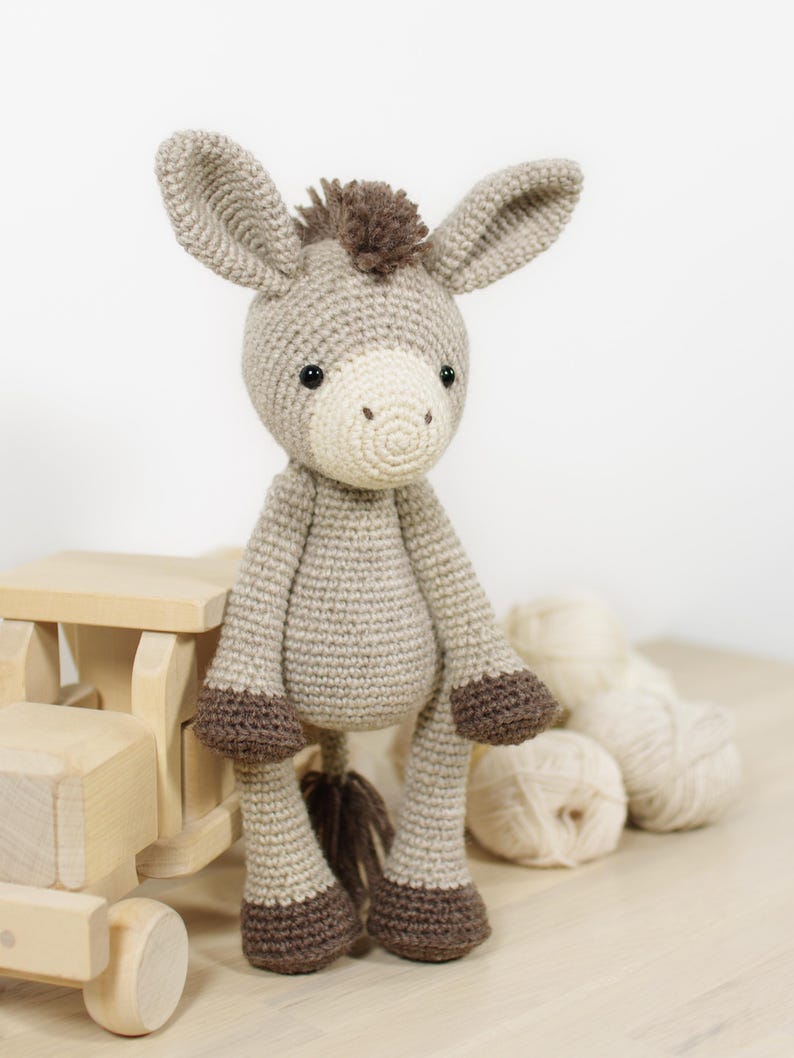May include: A crocheted donkey soft toy made from light beige yarn, featuring a cream-coloured muzzle, dark brown hooves, and a dark brown tuft of hair on its head and tail. The donkey has two large ears and small black eyes, standing upright.