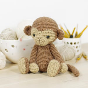 May include: A crocheted monkey stuffed animal featuring a dark brown body with a lighter tan face, ears, hands, and feet. The monkey has small black safety eyes and a stitched mouth detail. It is crafted with a dense, uniform yarn stitch pattern.