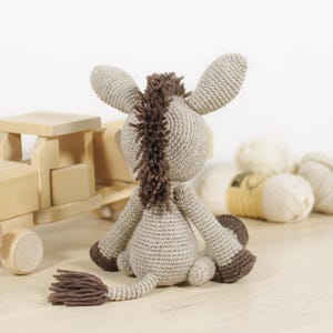 May include: A crocheted donkey toy in light beige with a dark brown mane, tail, and hooves. The donkey is seated, facing away from the viewer, showcasing its textured yarn construction, long ears, and a small tail with a tufted end.