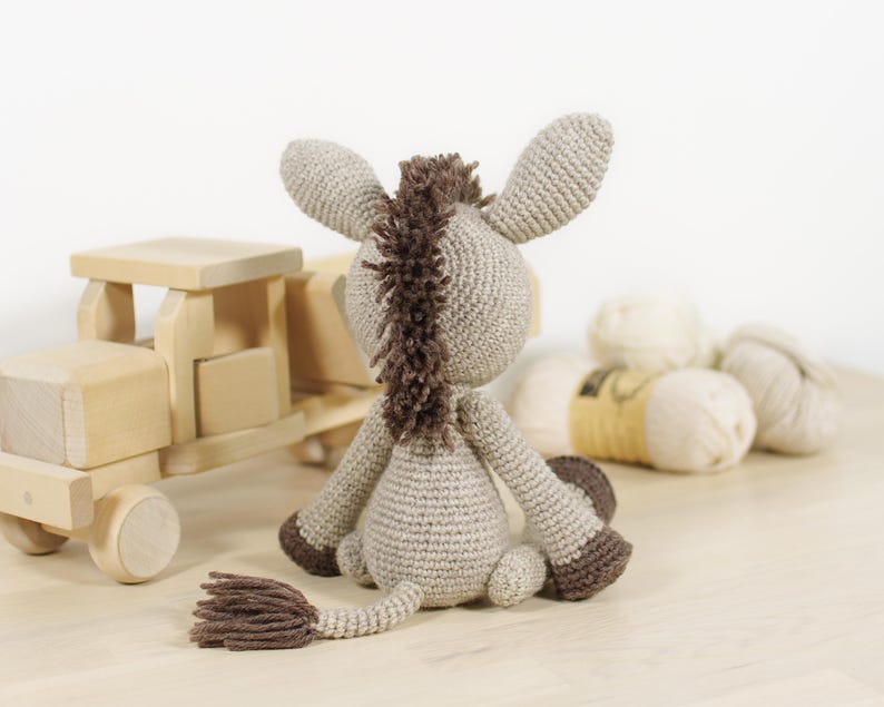 May include: A crocheted donkey toy in light beige with a dark brown mane, tail, and hooves. The donkey is seated, facing away from the viewer, showcasing its textured yarn construction, long ears, and a small tail with a tufted end.