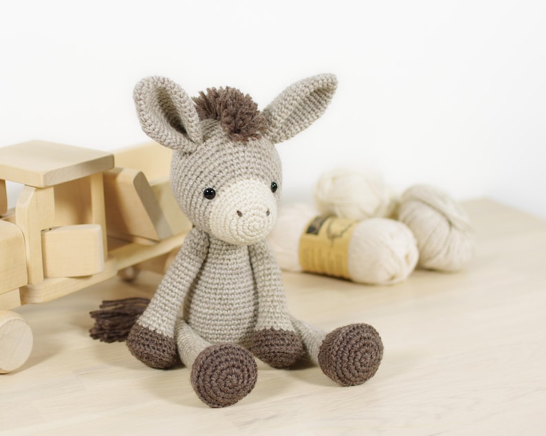 May include: A crocheted donkey toy with light grey fur, a cream-coloured muzzle, and dark brown hooves, mane, and tail. The donkey has two large, upright ears and small black eyes, sitting in an upright position with its legs extended forward.