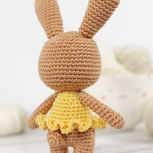 Small crocheted amigurumi bunny in a yellow ballerina outfit, standing in front of a yarn bowl and balls of yarn