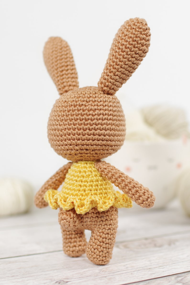 Small crocheted amigurumi bunny in a yellow ballerina outfit, standing in front of a yarn bowl and balls of yarn