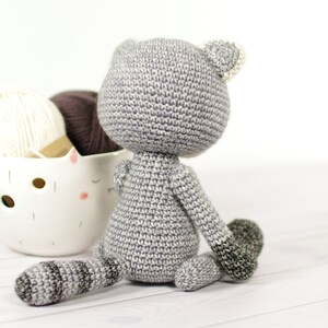 May include: A gray crocheted stuffed animal in the shape of a cat sitting on a white wooden surface. The cat has a white crocheted ear and a gray tail.