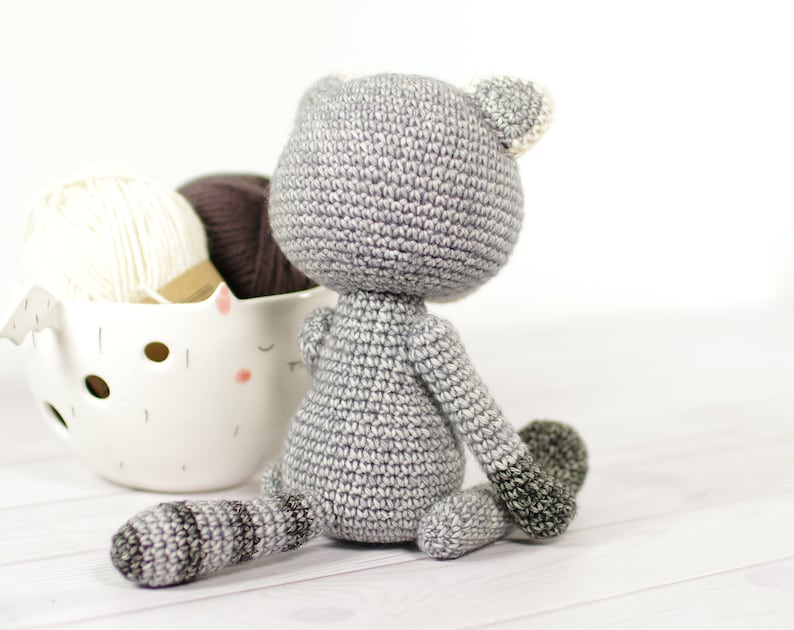 May include: A gray crocheted stuffed animal in the shape of a cat sitting on a white wooden surface. The cat has a white crocheted ear and a gray tail.