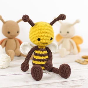 May include: A handmade crochet bee doll with a yellow head and brown and yellow striped body. The bee has white wings and brown legs and antennae. Two other crochet dolls are in the background.