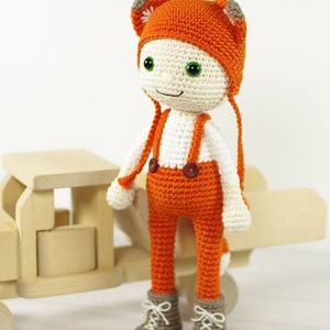 May include: A crocheted doll wearing an orange hat with fox ears, orange pants with suspenders, and brown boots. The doll has a white face with green eyes and a smile.