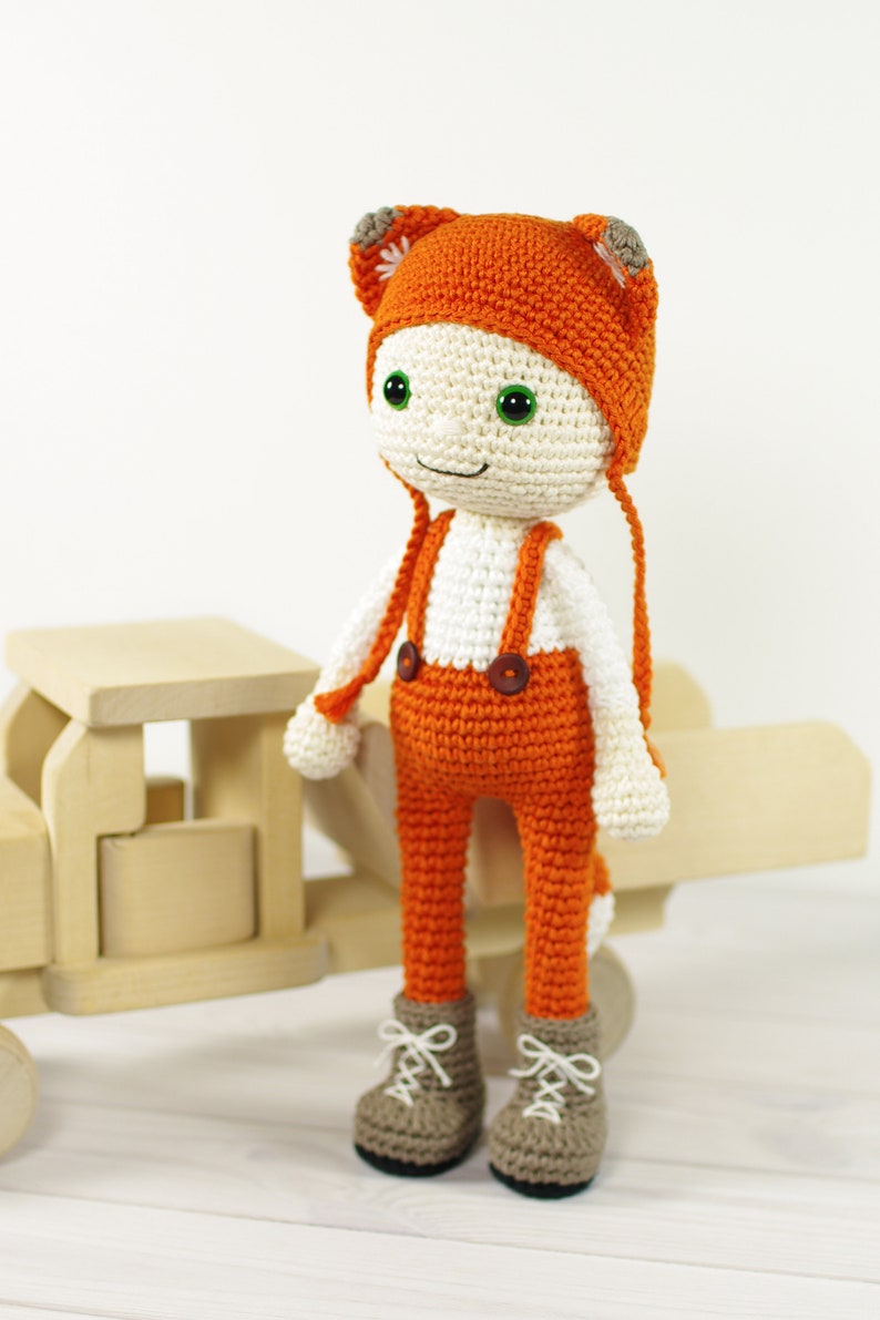 May include: A crocheted doll wearing an orange hat with fox ears, orange pants with suspenders, and brown boots. The doll has a white face with green eyes and a smile.