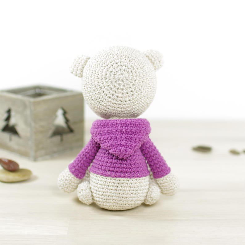 K&ouml;nnte beinhalten: Eine handgefertigte H&auml;kelb&auml;renpuppe, von hinten gesehen, mit einem lila Kapuzenpullover. Der B&auml;r ist haupts&auml;chlich cremefarben mit einem hellvioletten Pullover und wei&szlig;en Akzenten. Die Puppe steht auf einer hellen Holzoberfl&auml;che.