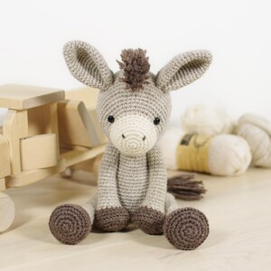 May include: A crocheted donkey toy with light beige yarn, a white muzzle, dark brown hooves, and a dark brown mane. The donkey has two large ears, black safety eyes, and a small tail. It is seated in an upright position with its legs extended forward.