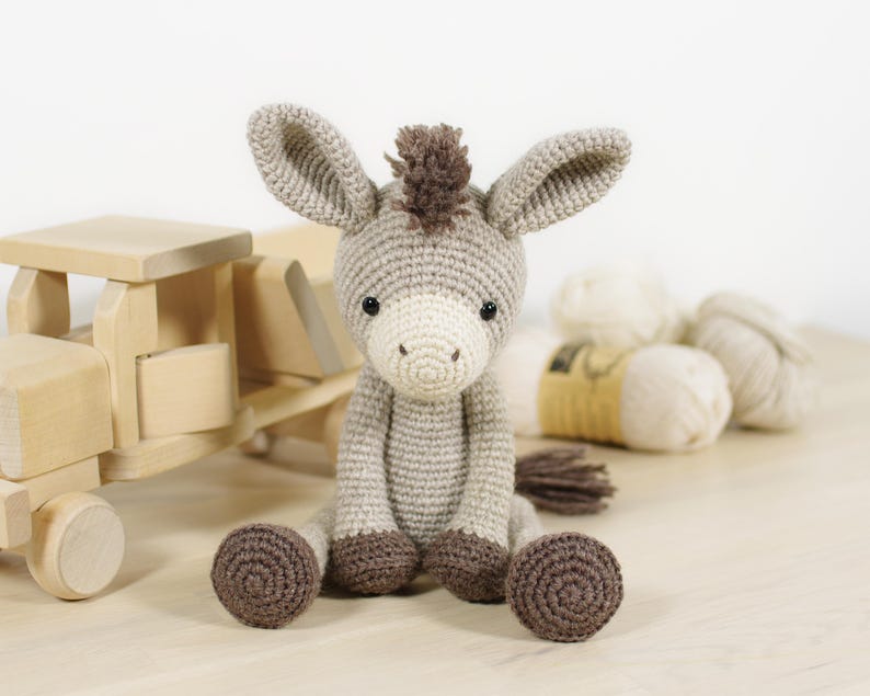 May include: A crocheted donkey toy with light beige yarn, a white muzzle, dark brown hooves, and a dark brown mane. The donkey has two large ears, black safety eyes, and a small tail. It is seated in an upright position with its legs extended forward.