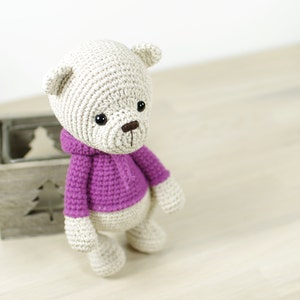 May include: A crocheted teddy bear wearing a purple hooded sweater. The bear has black eyes and a brown nose.