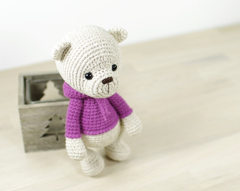 May include: A crocheted teddy bear wearing a purple hooded sweater. The bear has black eyes and a brown nose.