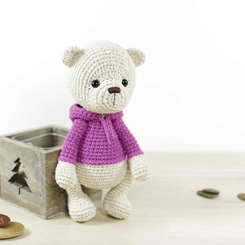 K&ouml;nnte beinhalten: Geh&auml;kelter Teddyb&auml;r in Beige mit lila Kapuzenpullover. Der B&auml;r hat eine braune Nase und schwarze Augen. Er sitzt neben einer Holzkiste mit einem Weihnachtsbaum-Design.