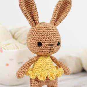 Small crocheted amigurumi bunny in a yellow ballerina outfit, standing in front of a yarn bowl and balls of yarn