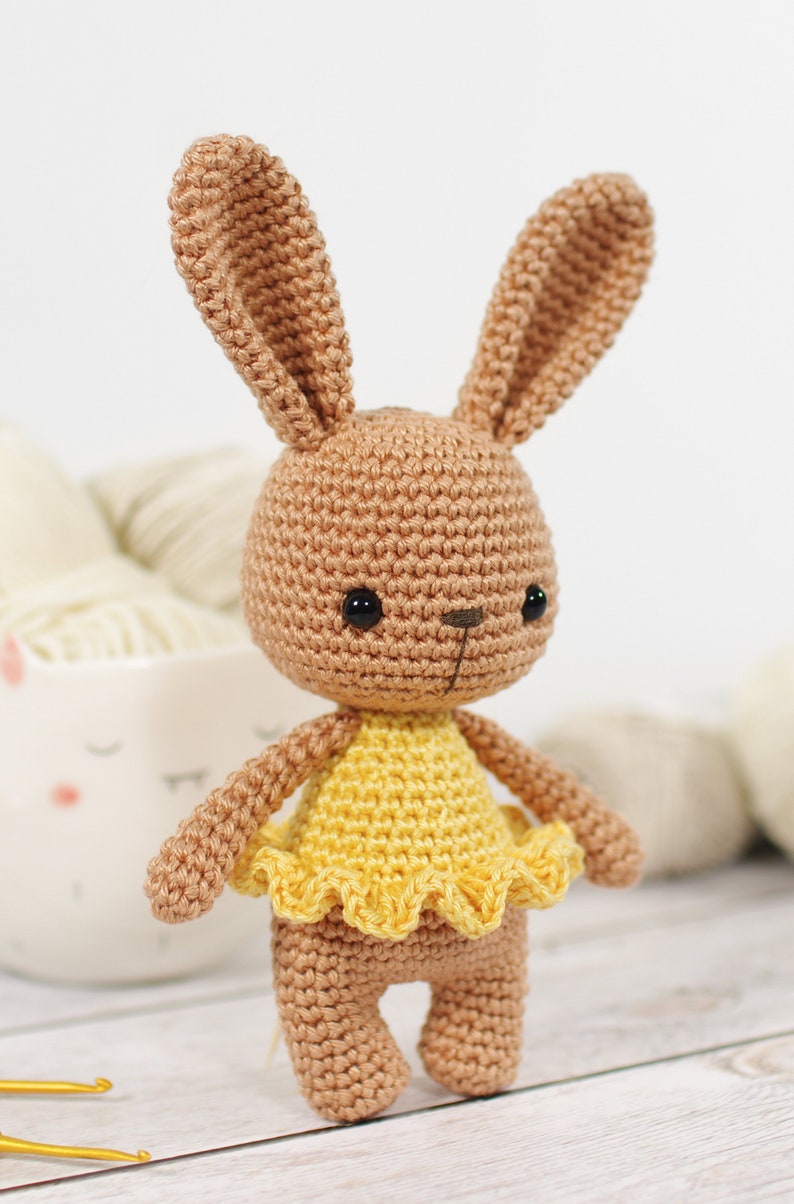 Small crocheted amigurumi bunny in a yellow ballerina outfit, standing in front of a yarn bowl and balls of yarn