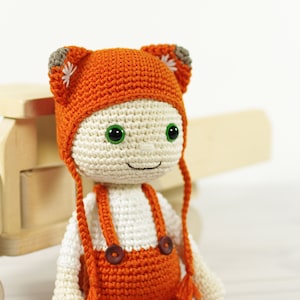 May include: A crocheted doll wearing an orange hat and overalls. The doll has green eyes and a smiling face. The doll is sitting on a wooden toy truck.