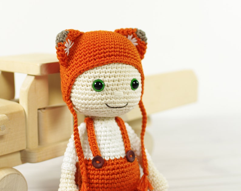 May include: A crocheted doll wearing an orange hat and overalls. The doll has green eyes and a smiling face. The doll is sitting on a wooden toy truck.