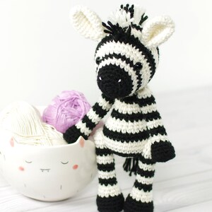 K&ouml;nnte beinhalten: Ein gestricktes Zebra-Spielzeug mit schwarz-wei&szlig;en Streifen, schwarzen Augen und einer schwarzen Nase. Das Zebra sitzt auf einer wei&szlig;en Holzoberfl&auml;che neben einer wei&szlig;en Keramikschale mit einem Comic-Gesicht.