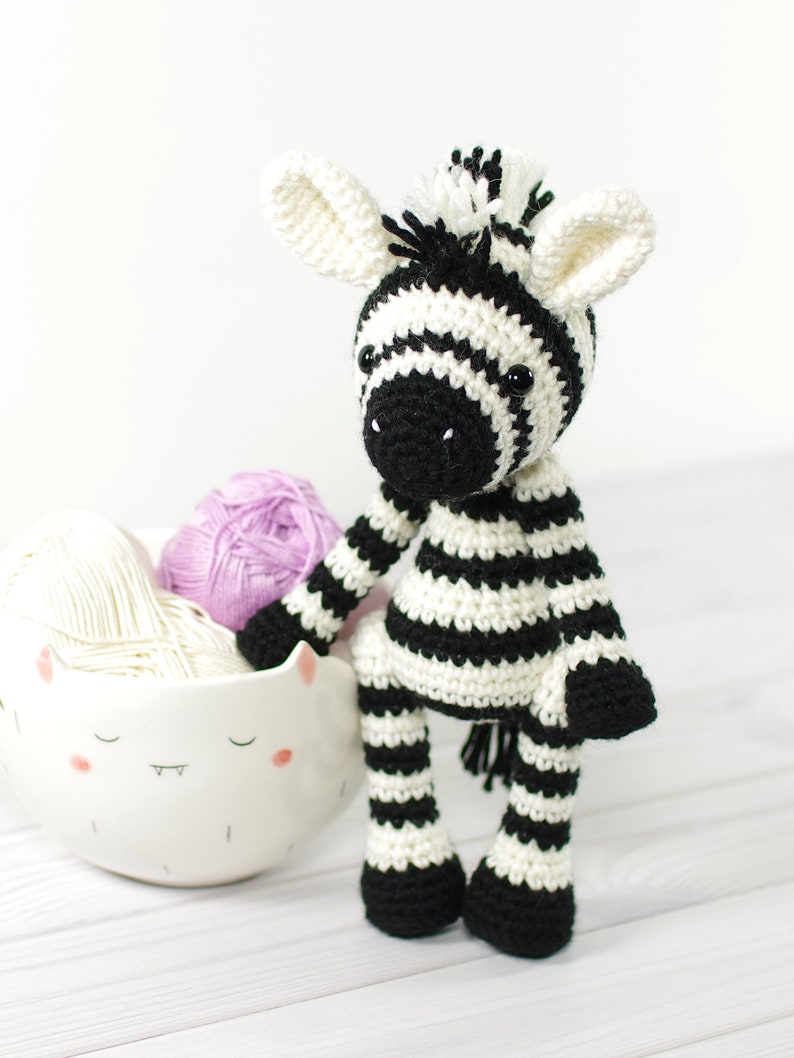 K&ouml;nnte beinhalten: Ein gestricktes Zebra-Spielzeug mit schwarz-wei&szlig;en Streifen, schwarzen Augen und einer schwarzen Nase. Das Zebra sitzt auf einer wei&szlig;en Holzoberfl&auml;che neben einer wei&szlig;en Keramikschale mit einem Comic-Gesicht.