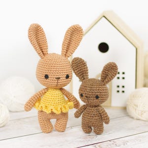 May include: Two crocheted bunny rabbits. One is larger, in tan with a yellow dress. The smaller bunny is brown. Both have black eyes and long ears. They are standing on a light wood surface, with a white house-shaped decoration in the background.