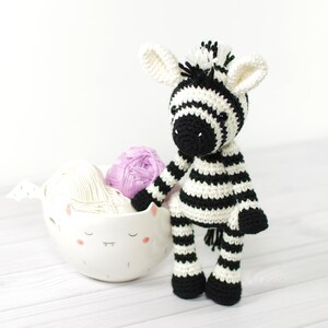 K&ouml;nnte beinhalten: Ein gestricktes Zebra-Spielzeug mit schwarzen und wei&szlig;en Streifen vor einem wei&szlig;en Hintergrund. Das Zebra steht auf einer wei&szlig;en Oberfl&auml;che mit einer wei&szlig;en Keramikschale gef&uuml;llt mit Garn.